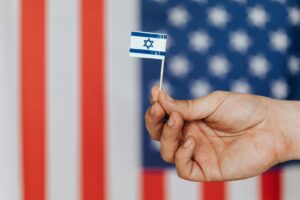 Close-up of a hand holding a small Israeli flag with American flag blurred in the background.