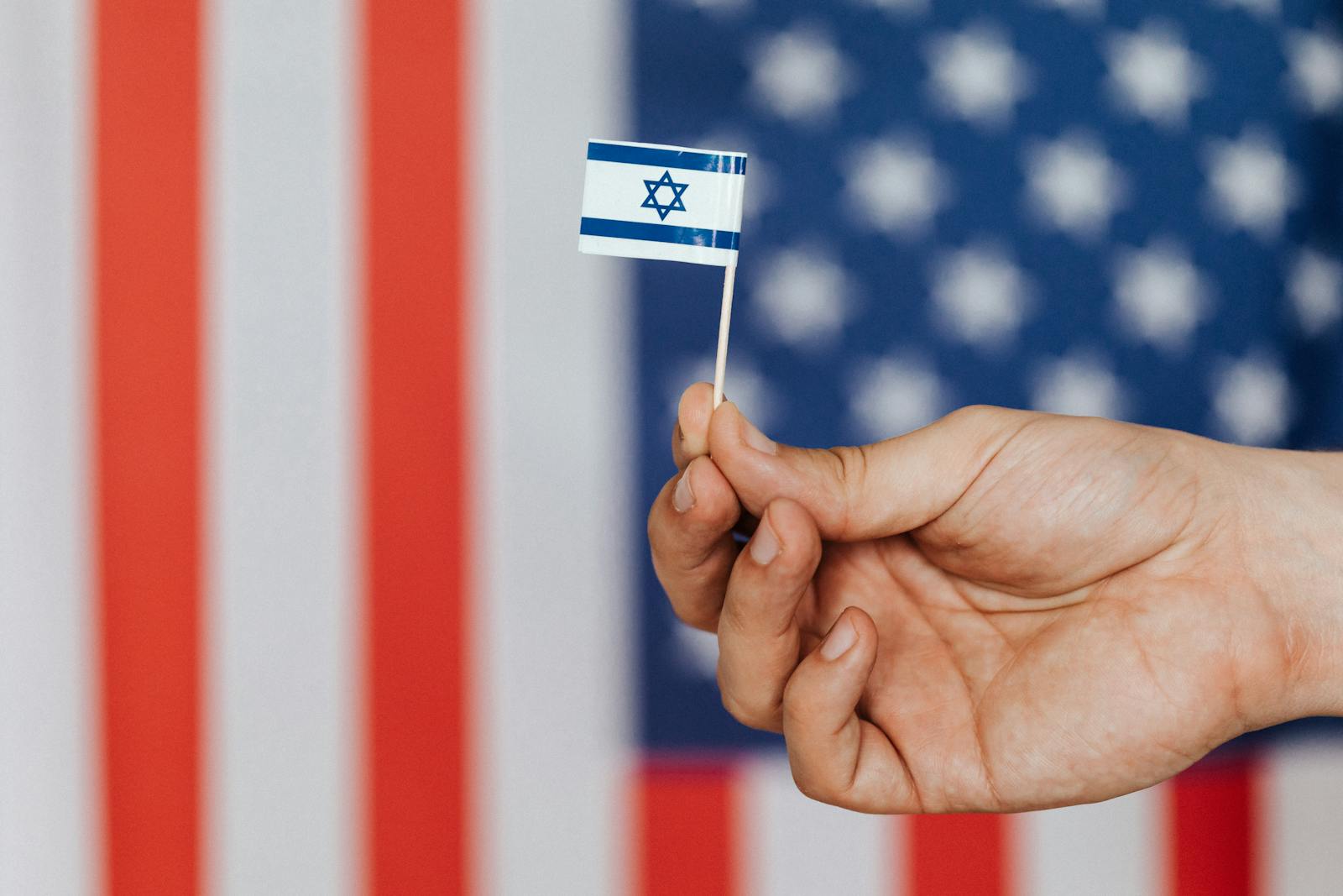 Close-up of a hand holding a small Israeli flag with American flag blurred in the background.