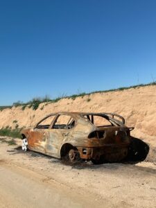 A rusty, burnt car wreck abandoned on a rural road against a dirt embankment under a clear blue sky.