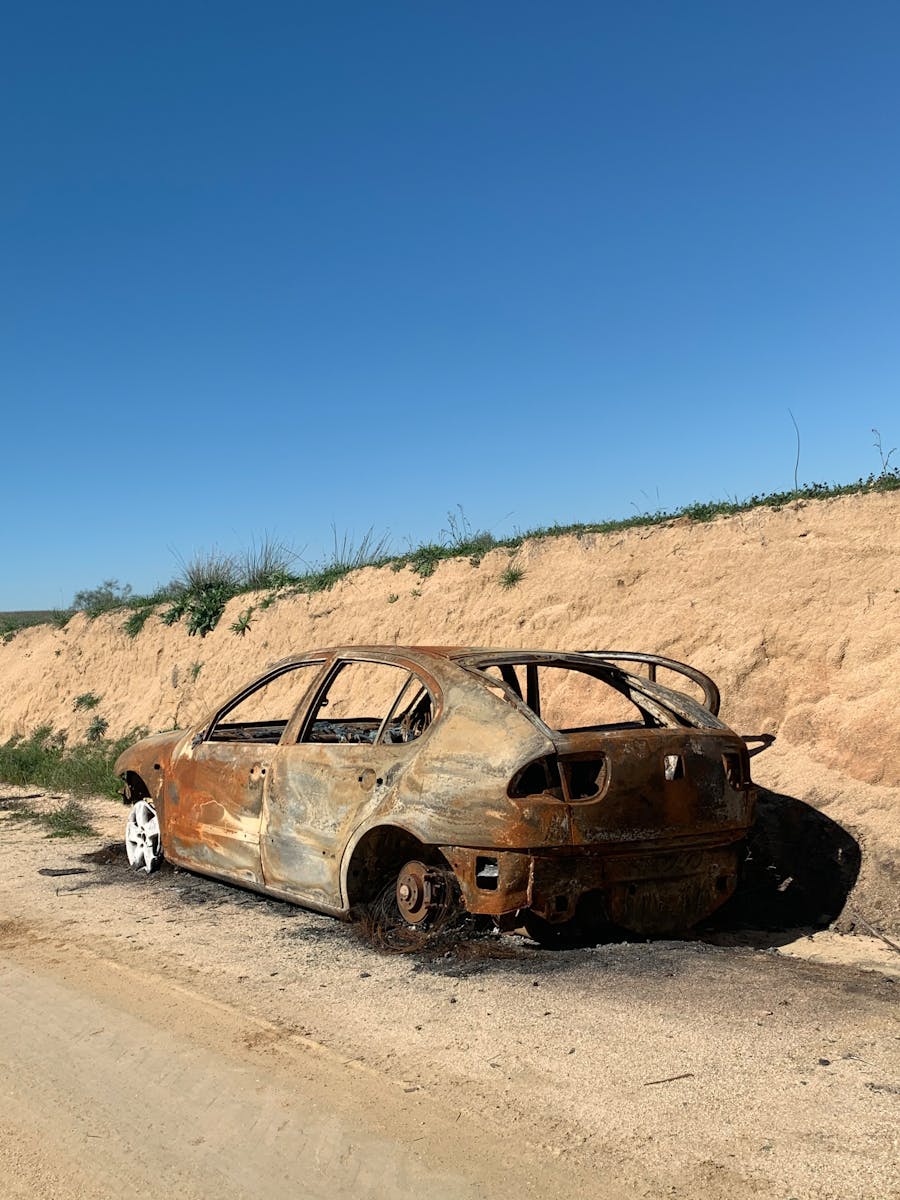 A rusty, burnt car wreck abandoned on a rural road against a dirt embankment under a clear blue sky.