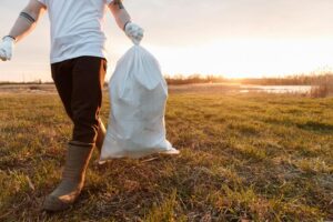 A volunteer carrying a garbage bag during a clean-up in a sunny outdoor field.