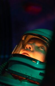 A female astronaut in a space suit looks up, illuminated by cosmic light.