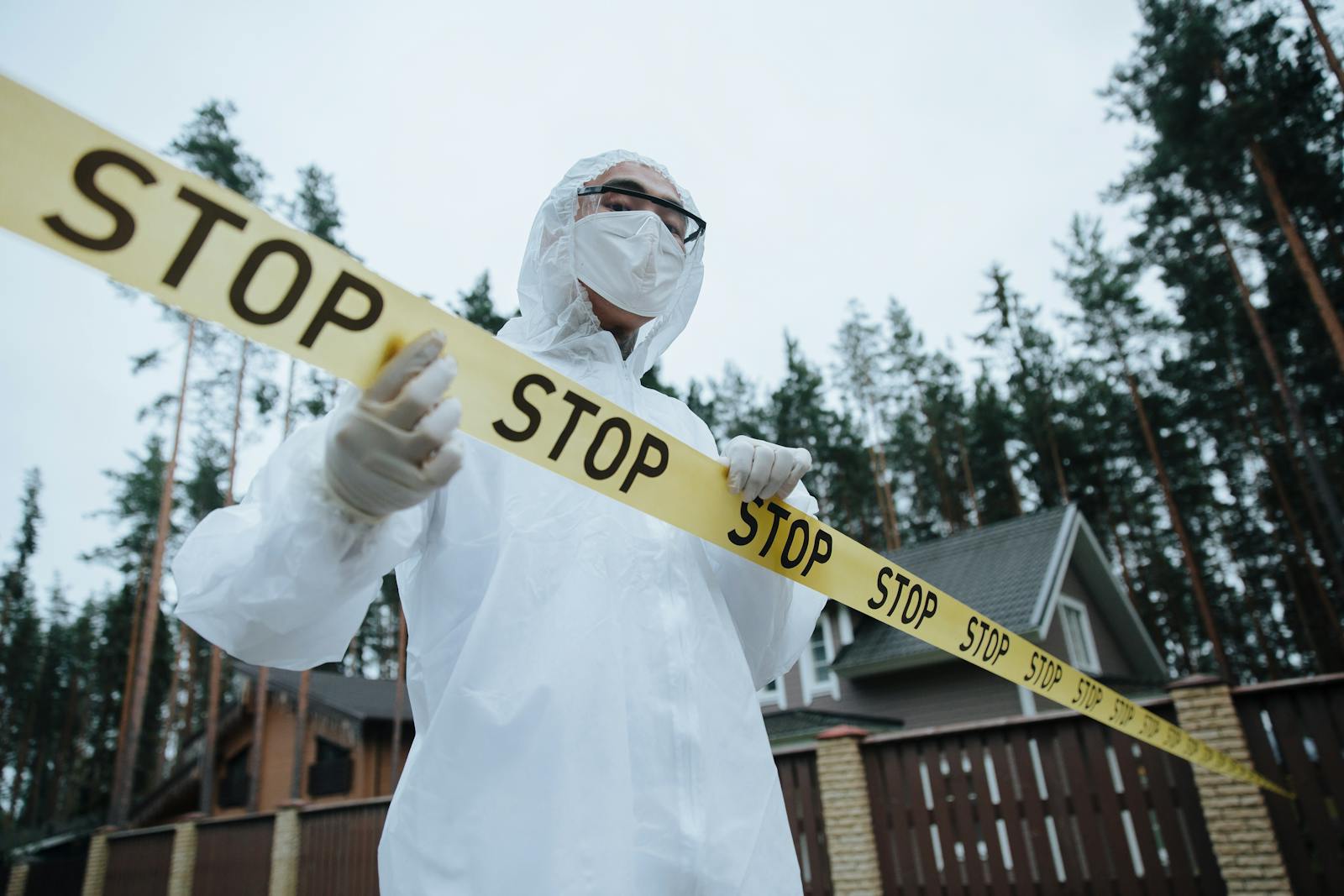 A forensic investigator in protective gear handles caution tape by a house in a wooded area.