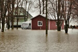 House and caravan surrounded by floodwaters among leafless trees in a natural setting.
