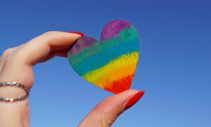 A hand holding a rainbow-colored heart against a clear blue sky, symbolizing love and pride.
