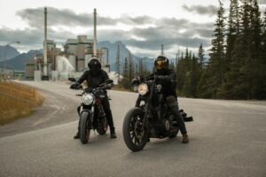 Two motorcyclists pause on a road with a factory and forest backdrop in Canada.