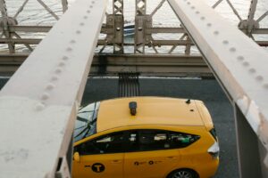 A New York City yellow taxi crossing the iconic Brooklyn Bridge over a river.