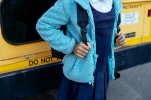 A schoolgirl in a blue jacket standing near a yellow school bus with a backpack.