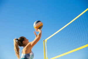 A woman plays volleyball on a sunny day, reaching to hit the ball over the net.