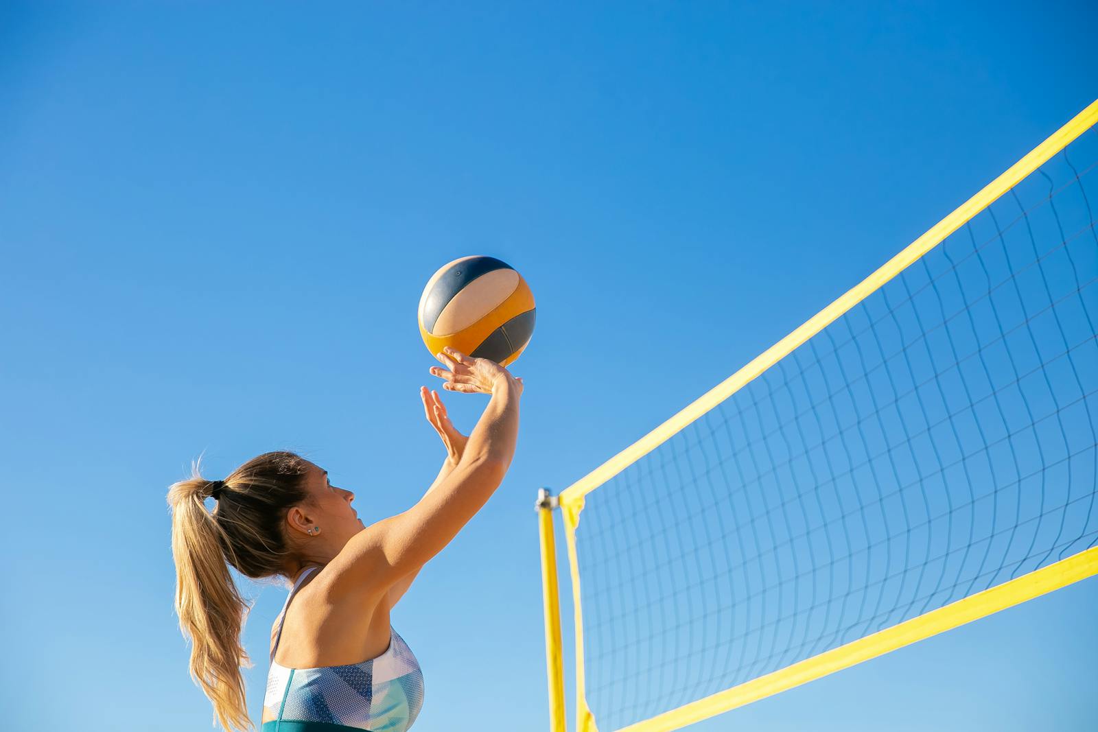 A woman plays volleyball on a sunny day, reaching to hit the ball over the net.