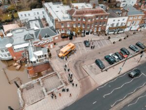 Drone view of school building facade near asphalt roadway and parking area with cars and bus with sidewalk and people near river in city street in daytime