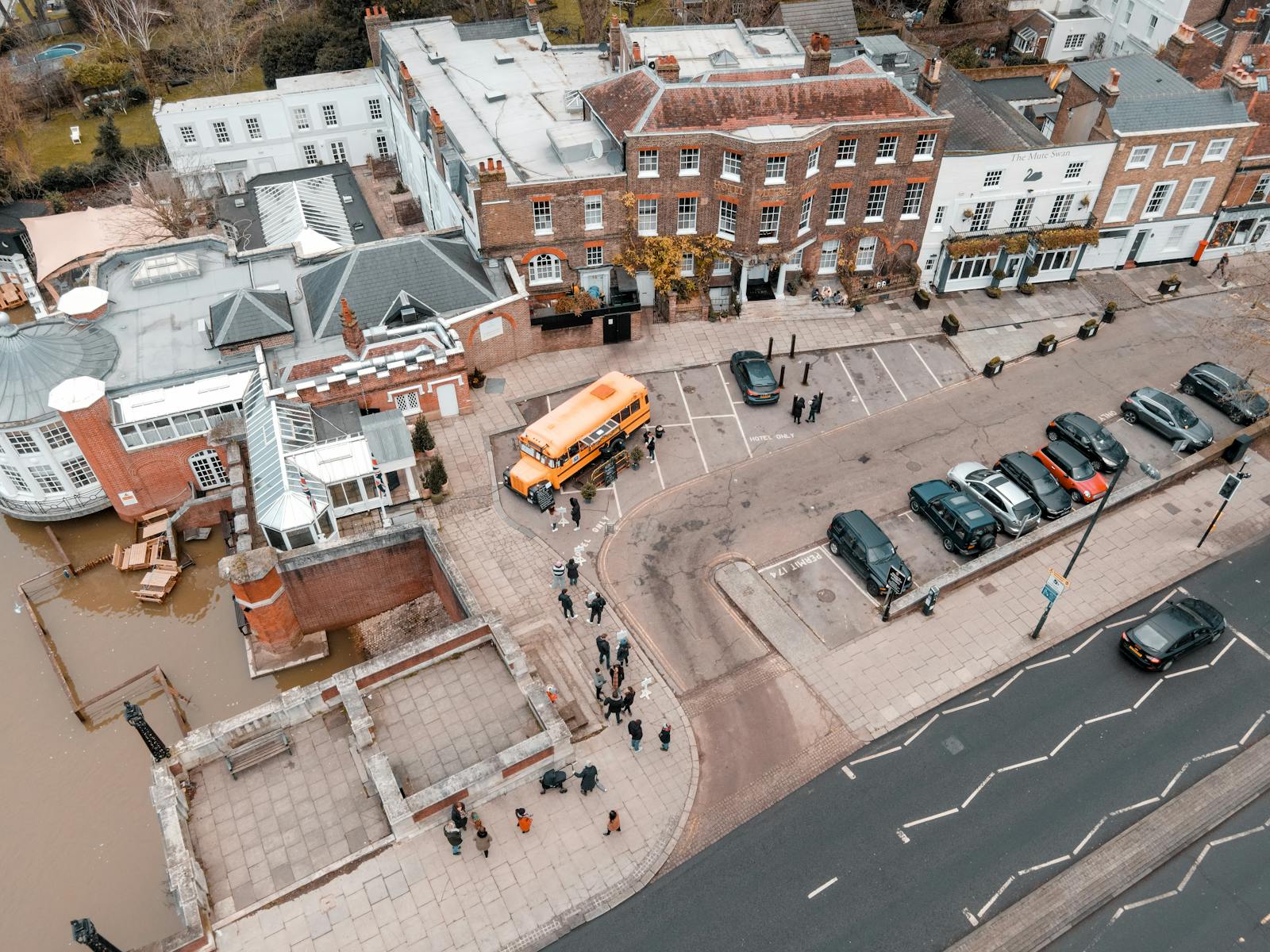 Drone view of school building facade near asphalt roadway and parking area with cars and bus with sidewalk and people near river in city street in daytime