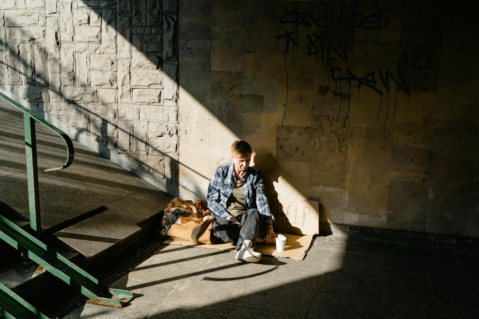 A homeless person sitting in a sunlit urban area, highlighting social issues.
