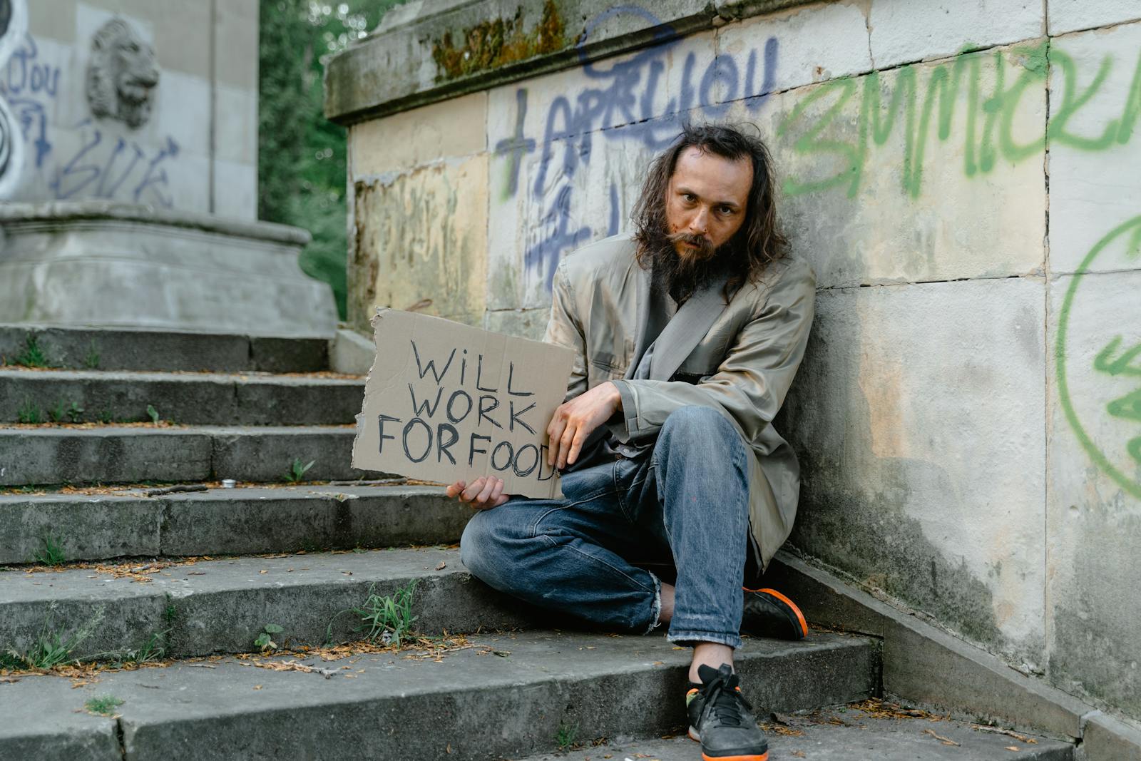 A homeless man sits on steps outdoors holding a 'Will Work for Food' sign, depicting poverty.