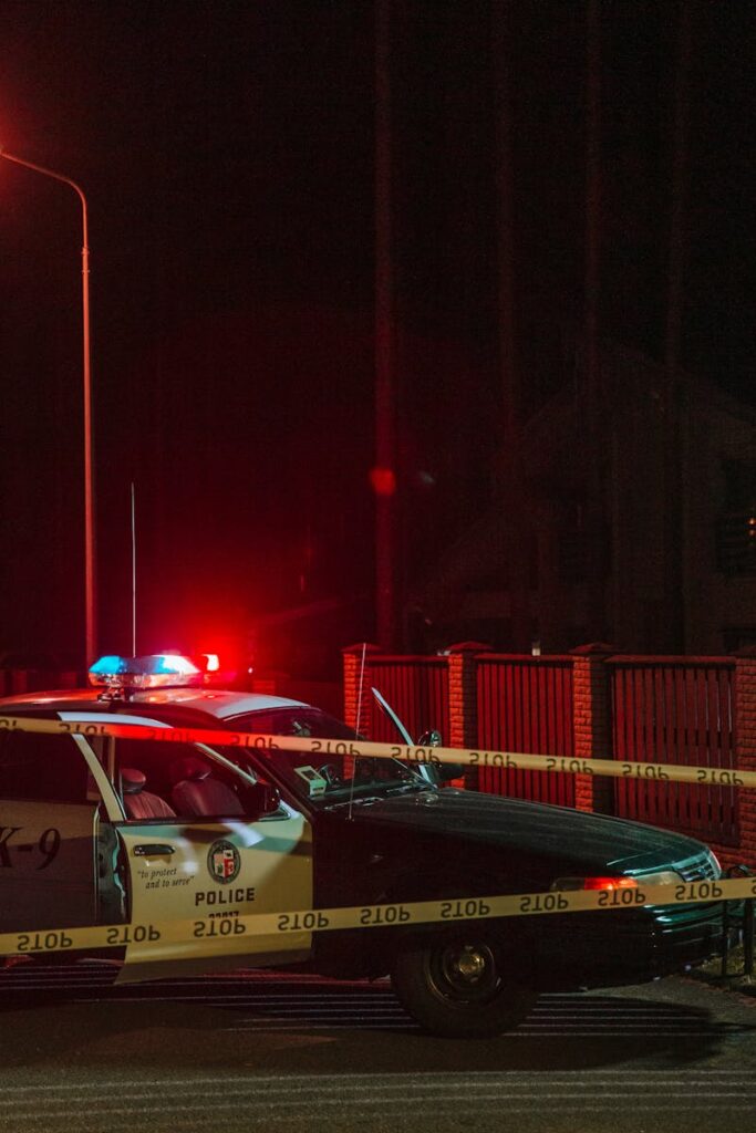 A police car with flashing lights at a nighttime crime scene secured with caution tape.